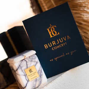 Pode incluir: Um frasco de vela de mármore branco com uma tampa e um rótulo dourados que dizem "Burjuva Concept Marble Series". O frasco de vela está sobre uma superfície branca ao lado de um cartão azul escuro com letras douradas que dizem "Burjuva Concept as special as you".