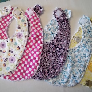 May include: Five baby bibs with different patterns. The bibs are made of soft fabric and have a white velcro closure. The patterns include floral, polka dots, zebra print, blue and white floral, and yellow with a giraffe and lion print.