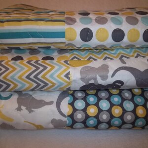 May include: A stack of three baby quilts with different patterns. The top quilt has a yellow, gray, and blue polka dot pattern. The middle quilt has a chevron pattern in yellow, gray, and blue. The bottom quilt has a white background with gray and yellow animal silhouettes.