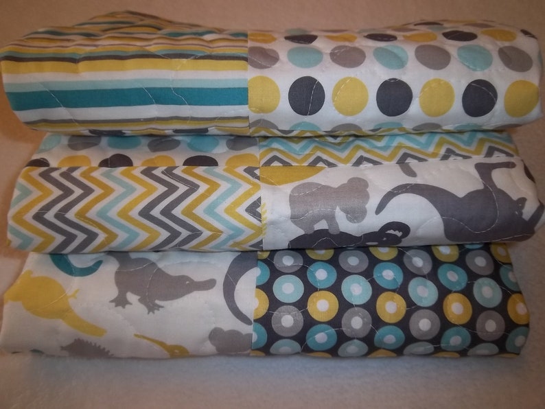 May include: A stack of three baby quilts with different patterns. The top quilt has a yellow, gray, and blue polka dot pattern. The middle quilt has a chevron pattern in yellow, gray, and blue. The bottom quilt has a white background with gray and yellow animal silhouettes.