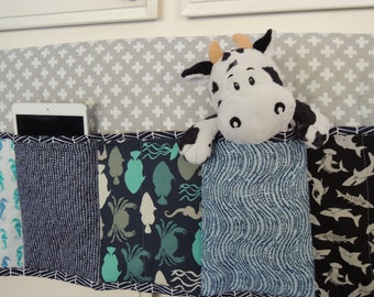 May include: A fabric bed organizer with multiple pockets featuring marine life patterns, including seahorses, fish, and sharks. A stuffed cow toy rests on top. The organizer has a gray and white cross pattern on the top and holds a tablet.