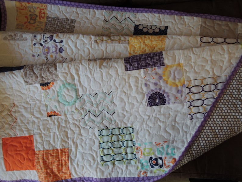 May include: A white quilt with a purple border and a patchwork design. The quilt is made up of squares of different fabrics in various colors, including orange, yellow, green, and purple. The quilt is folded in half and the back side is visible, which is a brown fabric with white polka dots.