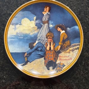 May include: A blue and white porcelain plate with a gold trim features a painting of four women sitting on a beach with a blue sky and white clouds in the background. The painting is signed "Norman Rockwell".