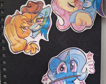 Può includere: Tre adesivi colorati con pony dei cartoni animati. Un adesivo mostra due pony che si abbracciano, un altro mostra due pony che si guardano e il terzo mostra un pony blu con una bacchetta e delle stelle.