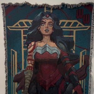 May include: Woven throw blanket featuring a stylized illustration of Wonder Woman in a red and blue costume. The artwork includes a teal background with gold accents and the Wonder Woman logo. The blanket has a fringed edge.
