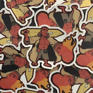 Puede incluir: Una colección de pegatinas troqueladas que representan a un personaje rojo y con cuernos con una gabardina beige, sosteniendo una gran espada amarilla. Las pegatinas tienen un borde blanco y están esparcidas sobre una superficie oscura.