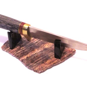 May include: A knife with a wooden handle and a silver blade is displayed on a stand made of a brown and white patterned stone. The stand has two black plastic supports.