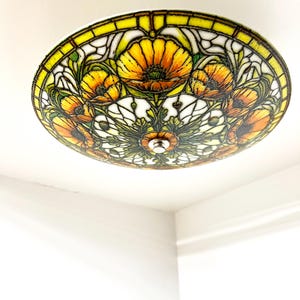 May include: A round ceiling light fixture with a stained glass design. The fixture features a floral pattern with yellow, orange, and green colors. The light is mounted on a white ceiling.