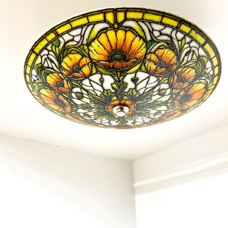 May include: A round ceiling light fixture with a stained glass design. The fixture features a floral pattern with yellow, orange, and green colors. The light is mounted on a white ceiling.