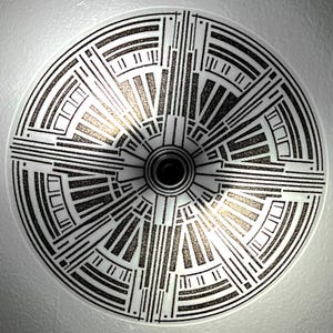 May include: A circular ceiling light with an Art Deco design. It has a white base with a black geometric pattern, including lines and rectangles, emanating from a central black cylinder. The centre has a glittery gold-toned finish.