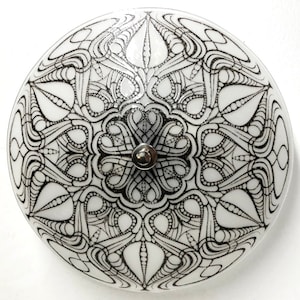 May include: A white glass ceiling light fixture with a black and white intricate design. The design is a mandala pattern with a central point in the middle.