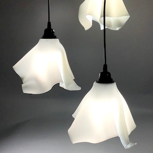 May include: Three white, fabric pendant lights with black cords hanging from the ceiling. The lights are shaped like a flower with ruffled edges.