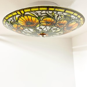May include: A round, stained glass ceiling light fixture with a floral design. The fixture features yellow, orange, and green flowers with a white and black background. The edge of the fixture is yellow and the base is white.