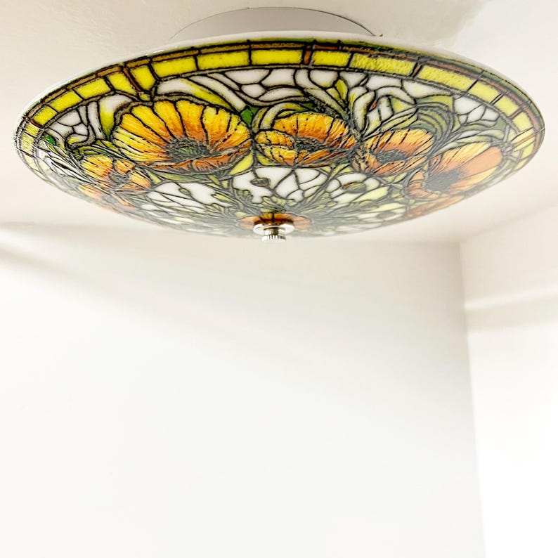 May include: A round, stained glass ceiling light fixture with a floral design. The fixture features yellow, orange, and green flowers with a white and black background. The edge of the fixture is yellow and the base is white.