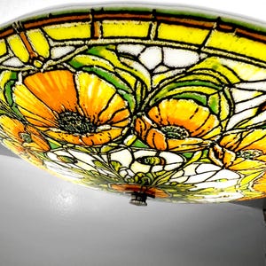 May include: A stained glass ceiling light fixture featuring a floral design with orange and yellow poppies. The flowers are set against a white and green background, with black outlines. The fixture has a metal base.