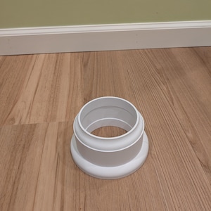 May include: A white plastic plumbing fitting with a circular base and a cylindrical top. The fitting has a stepped design, with a wider base and a narrower top. It is sitting on a wooden floor.