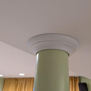 May include: A light green cylindrical column with a white circular trim at the top, attached to a white ceiling. The column is smooth and glossy. The background includes a yellow curtain and a black speaker.
