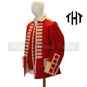 Op de afbeelding: Een rode jas in militaire stijl met witte biezen en zilveren knopen. De jas heeft een hoge kraag en decoratieve manchetten. De tekst "The Highland Tailor" is zichtbaar op de voorkant van de jas.