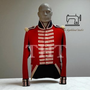 Op de afbeelding: Rode militaire jas met wit en goudkleurige details, gepresenteerd op een mannequin. De jas heeft een hoge kraag, meerdere knooprijen en epauletten. De tekst "the highland tailor" is zichtbaar.