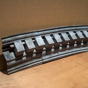 May include: A curved, gray, metal architectural molding with a repeating pattern of rectangular blocks. The molding is made of a textured material and has a slightly weathered appearance.