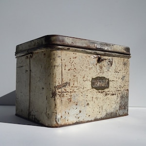 May include: A vintage, white metal tin box with a rusted lid and a label that reads "Premier". The box has a worn, distressed look and is likely used for storage.