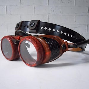May include: Red and black steampunk goggles with a black leather headband. The goggles have a clear lens and the headband has a metal buckle.