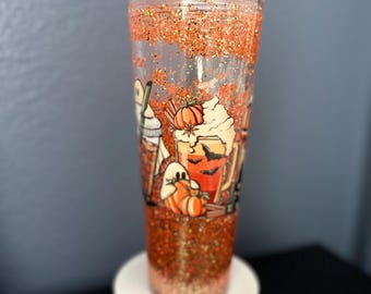 May include: A clear, double-walled tumbler filled with orange glitter and a Halloween-themed design. The design includes pumpkins, bats, ghosts, and a coffee cup. The tumbler is on a white stand.