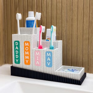May include: A multi-tiered toothbrush holder with the words "Daddy", "Mommy", "Mia", and "May" in colorful letters. The holder contains toothbrushes, toothpaste, and floss. The base is black, and the holder is a light gray color.