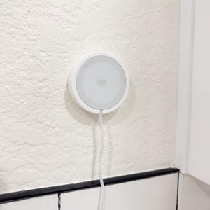 May include: A white, circular Qi wireless charger mounted on a textured white wall. The charger has a gray center with the Qi logo and a white cable extending downwards. The wall has a rough, stucco-like finish.