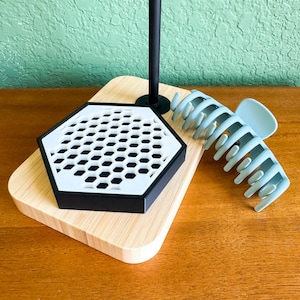 May include: A black and white hexagonal tray with a honeycomb pattern sits on a wooden base. A light blue plastic hair claw is placed next to the tray.