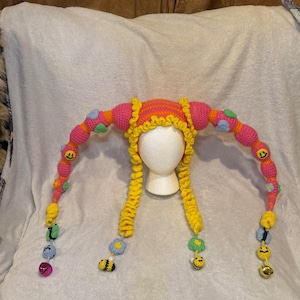 Op de afbeelding: Een grillige, handgemaakte gehaakte hoed in helder roze, geel en oranje. De hoed heeft lange, kralenranken met smiley-gezichten, bloemen en bijenaccenten. De hoed is ontworpen met een unieke, speelse esthetiek.