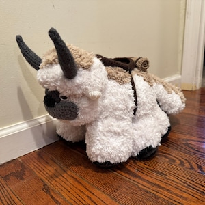 May include: A plush toy of a flying bison, featuring a white body with brown and tan accents. The toy has large, dark gray horns and a crocheted face. A brown saddle is attached to its back. The toy is sitting on a wooden floor.