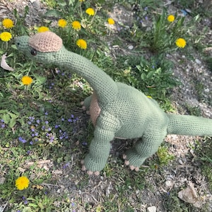 Puede incluir: Un juguete de dinosaurio de ganchillo verde hecho a mano con un cuello y cola largos, y un parche marrón en la espalda. El dinosaurio está posado entre hierba, dientes de león y pequeñas flores moradas.