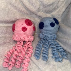 May include: Two handmade crochet jellyfish toys. One is pink with red spots and pink tentacles. The other is light blue with dark blue spots and light blue tentacles. The jellyfish are on a soft, white, textured surface.