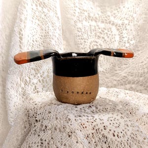 May include: A small, handmade ceramic ashtray. The ashtray has a brown base with small holes, a black upper section, and two wing-like extensions with orange and black accents. The ashtray is set against a white, lace-like background.