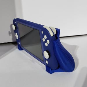 Puede incluir: Consola de juegos portátil azul con botones blancos y pantalla oscura. La consola está en una funda de agarre azul para mejorar la ergonomía y la estabilidad. La funda tiene un soporte incorporado para jugar sobre la mesa.
