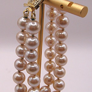 May include: A double-strand pearl bracelet with large, creamy-colored beads. The bracelet is held by a gold-toned clasp with intricate detailing. The pearls are displayed on a gold-colored stand.