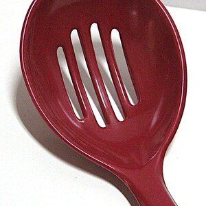 May include: A deep, dark red slotted spoon with a wide, rounded bowl. The spoon features four vertical, elongated slots centered in the bowl, designed for draining liquids while serving food. The surface has a smooth, glossy finish throughout.