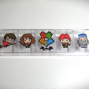 May include: A clear plastic container with five compartments, each featuring a different cartoon character from the Harry Potter series. The characters include Harry Potter on a broom, Hermione Granger, the Hogwarts crest, Ron Weasley, and Dumbledore.