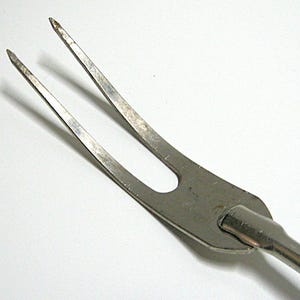 May include: Close-up of a vintage metal carving fork with two tines. The fork has a dull, silver finish and a long, slender handle. The tines are slightly curved and pointed. The background is plain white.