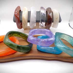May include: A collection of colorful, square-shaped bangles in various colors, including orange, green, purple, and blue. Some bangles are displayed on a wooden tray, while others are on a display stand. The bangles have a glossy finish.