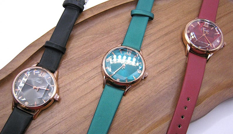May include: Three elegant wristwatches with rose gold cases and leather straps in black, teal, and burgundy. Each watch features a unique dial design and the brand name "Tadi". The watches are displayed on a wooden surface.