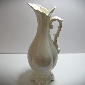 May include: A cream-colored ceramic pitcher with a decorative handle and a scalloped rim. The pitcher has a bulbous body and a detailed base. The handle is ornate, and the overall design is elegant.