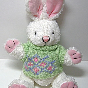May include: A plush white bunny with pink inner ears, nose, and paw pads. The bunny wears a light green sweater with a floral pattern in pink and blue. The bunny is a stuffed animal.