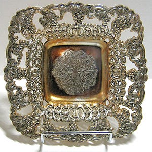 May include: Silver-toned square tray with an ornate grape vine design around the edges. The center of the tray has a decorative floral pattern. The tray is supported by a metal stand.