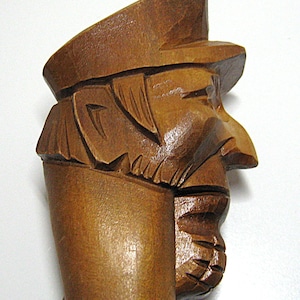 May include: A hand-carved wooden sculpture of a man wearing a hat. The sculpture is a medium brown color and features detailed carving of the man's face, including his nose, mouth, and beard. The hat has a flat top and a brim. The sculpture is likely a decorative item.