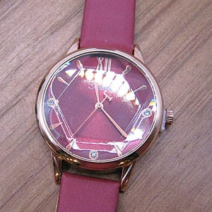 May include: A rose gold wristwatch with a burgundy face and band. The watch face features a geometric design with Roman numerals and small blue accents. The band is made of a smooth, burgundy material.