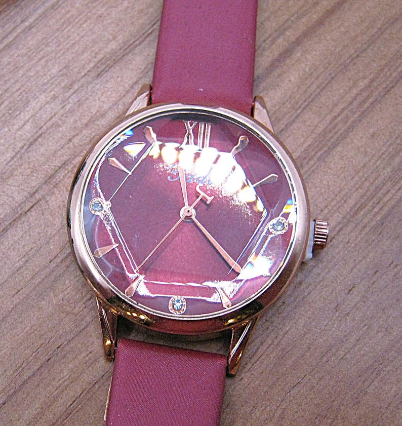 May include: A rose gold wristwatch with a burgundy face and band. The watch face features a geometric design with Roman numerals and small blue accents. The band is made of a smooth, burgundy material.