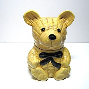 May include: A ceramic teddy bear figurine with a basket weave texture in a light tan color. The bear has a black bow tie and black eyes and nose. The ears are upright, and the bear is sitting.