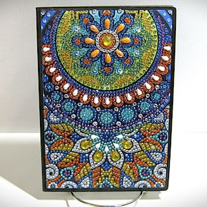 May include: A rectangular, colorful mandala art piece with a black border. The design features a symmetrical pattern of blue, green, orange, and yellow dots, creating a vibrant and detailed image. The artwork is displayed on a stand.